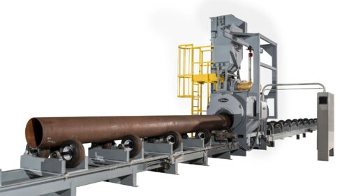 Image is of a large piece of machinery being fed a large cut of pipe to be blasted inside a wheel blast chamber. Machinery is grey photographed on a white background with a rusty pipe being rolled into it.