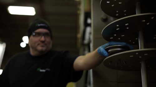 Image is of a SurfacePrep employee removing a finished part from the fixture inside a spinner hanger wheel blast system.