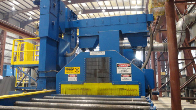 Image is of a piece of blue and yellow machinery with a large wheel blast corridor that is fed by a roll conveyor system. The machinery was photographed within a warehouse.